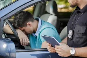 Shot of a young driver keeping his head on a steering wheel and a policeman standing next to the car